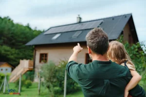 Un homme portant un enfant dans ses bras montre les panneaux solaires installés sur le toit d'une maison en bois, entourée de verdure et de nature.