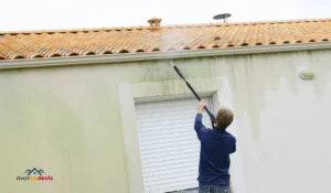 Un homme en train de nettoyer la façade d'une maison à l'aide d'un nettoyeur haute pression, enlevant des traces de saleté et de moisissure sur le mur sous le toit en tuiles.